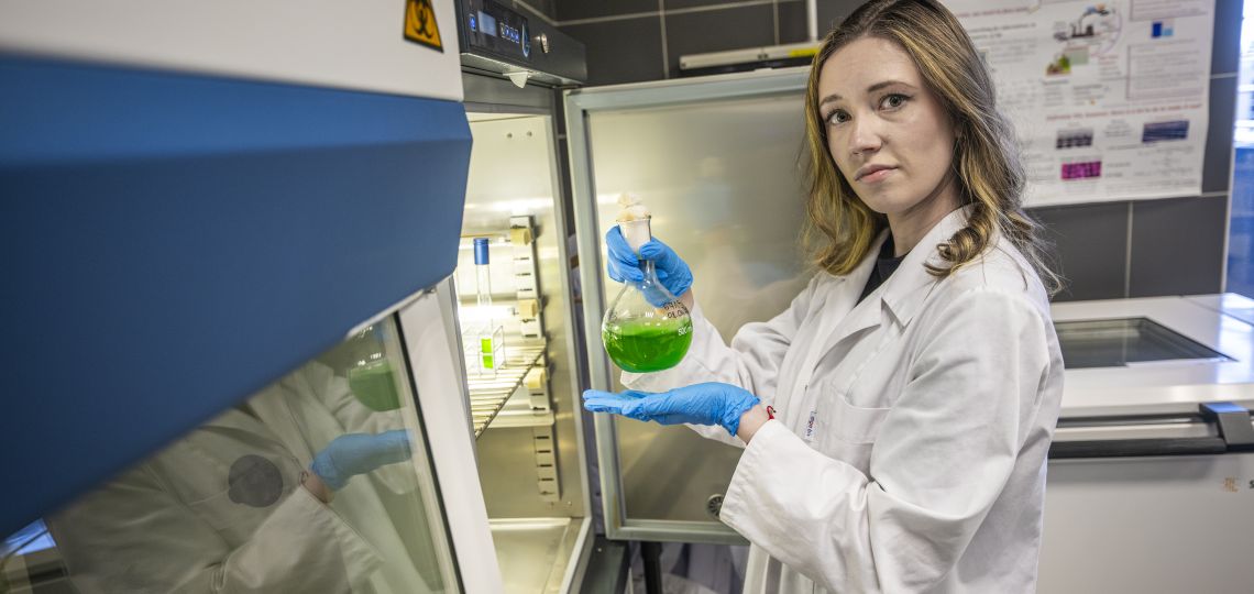 młoda kobieta w laboratorium, w fartuchu i niebieskich rękawiczkach ochronnych trzyma w dłoniach kolbę z zielonym płynem