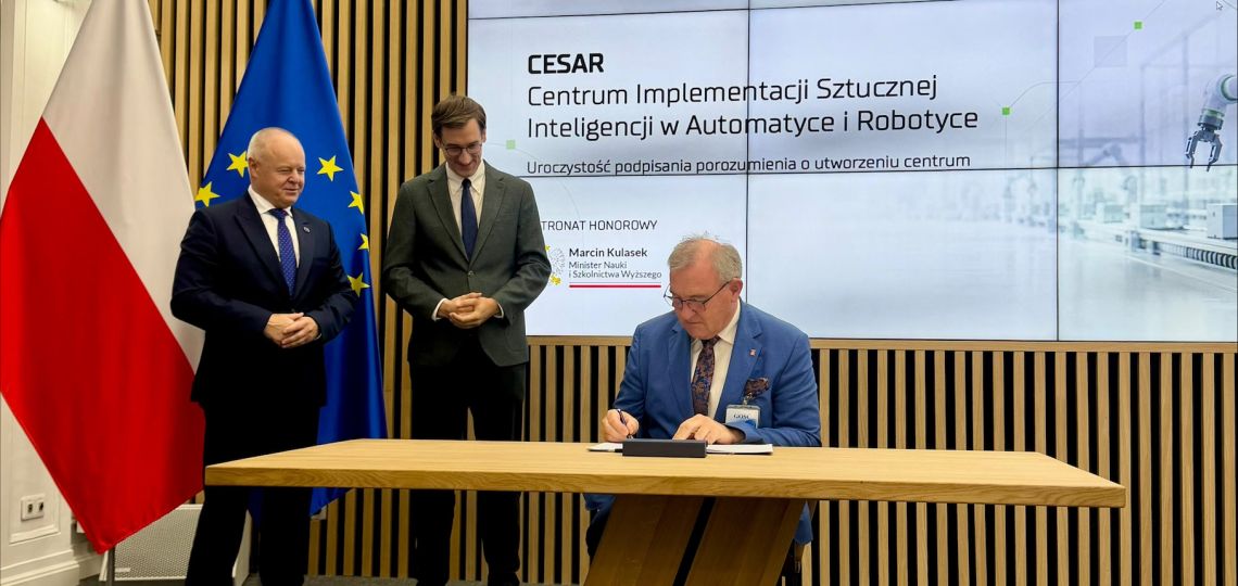 Uroczystość podpisania porozumienia o utworzeniu Centrum Implementacji Sztucznej Inteligencji w Automatyce i Robotyce (CESAR). Profesor Krzysztof Jóźwik siedzi przy drewnianym stole i podpisuje dokument, obok stoją dwaj mężczyźni w garniturach. W tle flagi Polski i Unii Europejskiej oraz ekran z logotypami Ministerstwa Nauki i Szkolnictwa Wyższego oraz Sieci Badawczej Łukasiewicz, a także napisem „CESAR – Centrum Implementacji Sztucznej Inteligencji w Automatyce i Robotyce – Uroczystość podpisania p