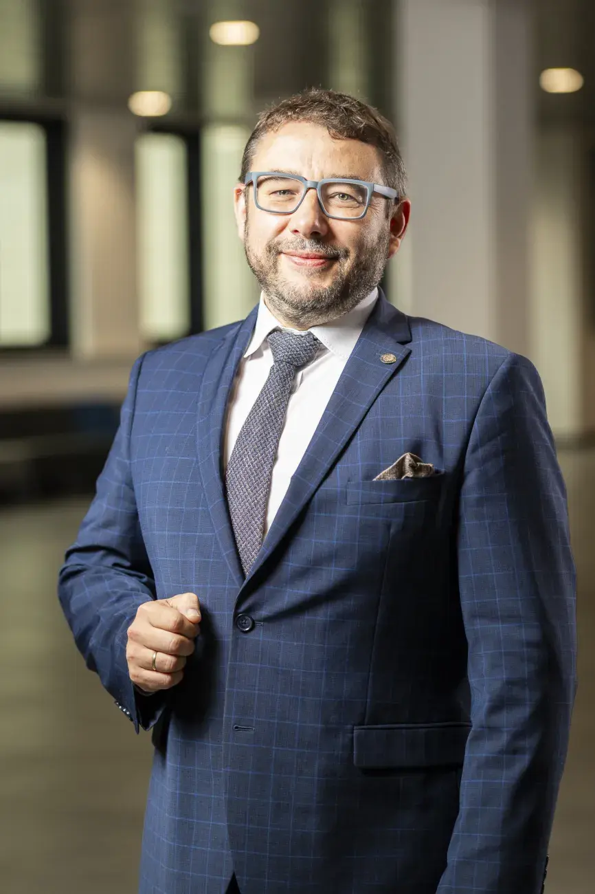 zdjęcie uśmiechniętego mężczyzny w garniturze i krawacie, w okularach, z brodą, stoi na tle przestrzennego korytarza