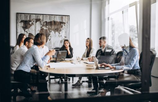Kilkanaście osób ubranych w oficjalne stroje siedzi w przeszklonej sali konferencyjnej przy okrągłym stole. W tle na ścianie mapa świata.