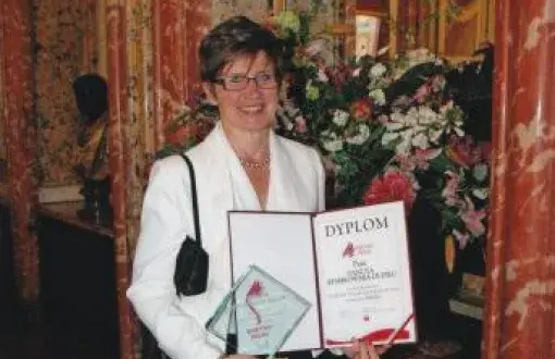 Danuta Kembłowska-Dupieu w białej marynarce trzyma w dłoniach i prezentuje dyplom i statuetkę. W tle elegancie wnętrze z kwiatami.