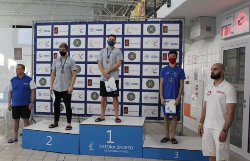 Niebieskie podium i na nim trzech mężczyzn w stronach sportowych. W tle ścianka z logami organizatorów.