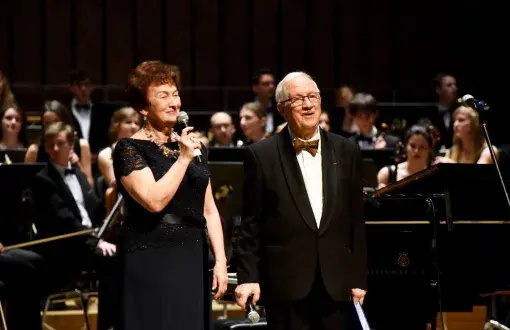 prof. Jan Krysiński w smokingu i Grażyna Sikorska w czarnej sukni na scenie Filharmonii Łódzkiej. W tle orkiestra.