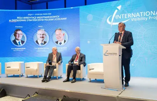 Jasna scena z 4 białymi fotelami i mównicą, przy której stoi prof. Krzysztof Jóźwik. W tle niebieska ścianka z napisami i zdjęciami panelistów.