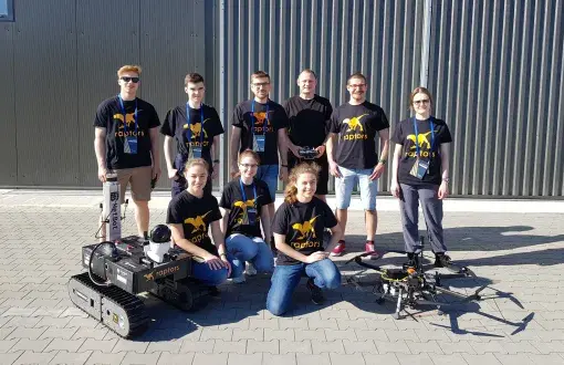 Członkowie drużyny Raptors z łazikiem i dronem na tle ciemnego budynku.