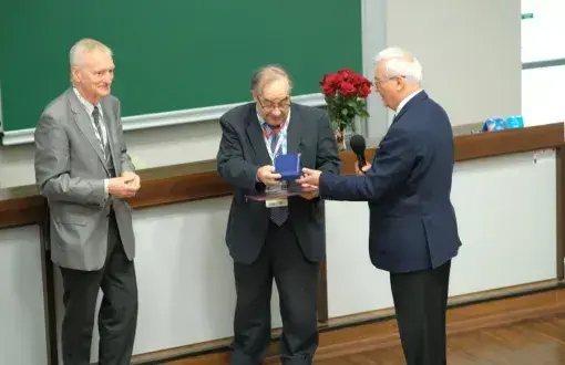 3 mężczyzn w starszym w sali wykładowej wręcza sobie medal i kwiaty