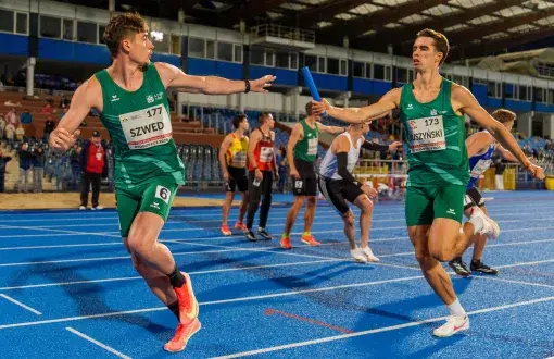 dwócg biegaczy w zielonych strojach przekazuje sobie pałeczkę w biegu, na stadionie