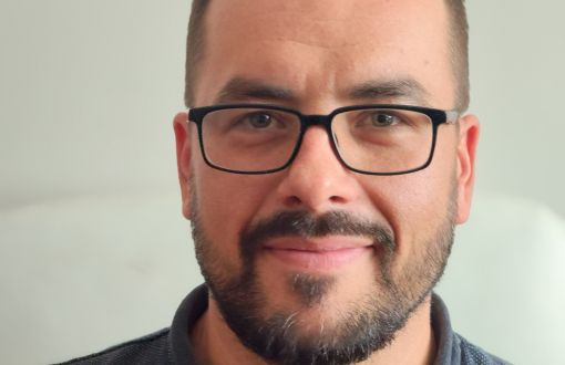 uśmiechnięty młody mężczyzna w okularach z czarnymi oprawkami stoi na tle beżowej ściany