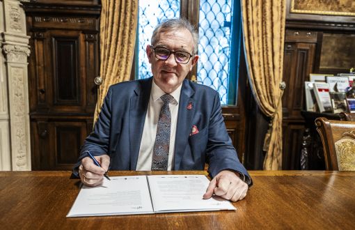 patrzący w obiektyw mężczyzna w okularach i garniturze siedzi przy stole i podpisuje dokumenty piórem