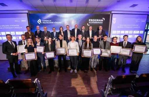 grupa ludzi na podświetlanej na fioletowo ścianie, w pierszym rzędzie młodzi ludzie trzymają dyplomy oprawione w złote ramki, w drugim kilkuosobowa grupa dojrzałych ludzi
