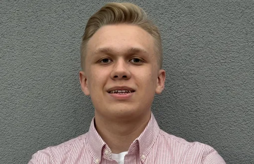portret młodego mężczyzny o blond włosach, opierającego się o szarą ścianę, ma na sobie koszulę w biało-różowe pionowe paseczki
