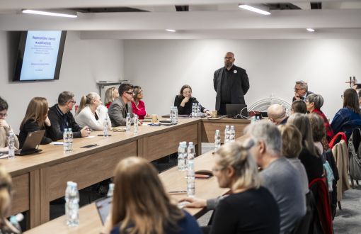 grupa osób siedzi przy ustawionych na przeciw siebie stołach, 1 mężczyzna stoi, jest ubrany na czarno