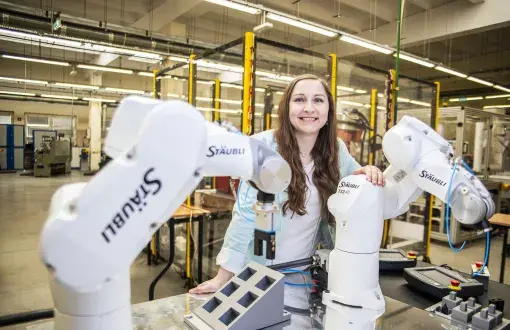 kobieta z długimi włosami nachyla się nad urządzeniem robotycznym, w tle wyposażenie hali technologicznej