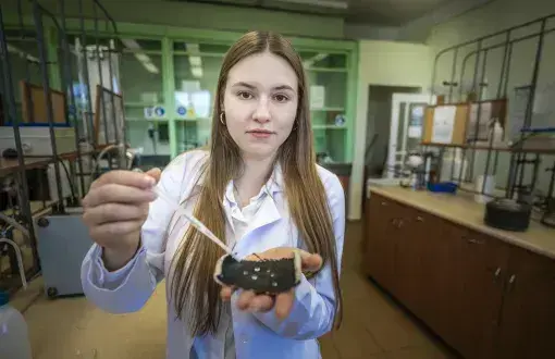 studentka w białym fartuchu trzyma pipetę oraz kawałek ciemnego materiału, na którym widać skroploną wodę, w tle zyblurowane laboratorium