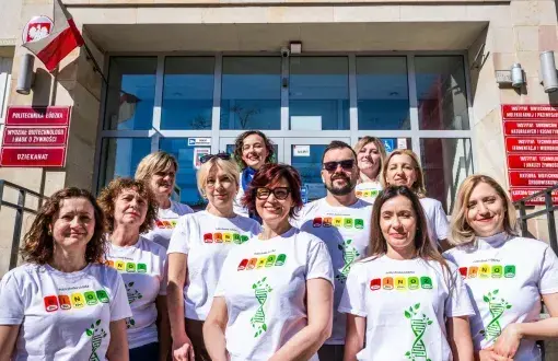 11 uśmiechniętych osób w białych koszulkach stoi na tle budynku z beżową fasadą i czewonymi tabliczkami informacyjnymi