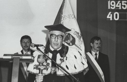 czarno-białe zdjęcie starszego mężczyzny w tradycyjnym stroju rektorskim, trzymającego berło, w tle sztandar trzymany przez dwóch mężczyzn