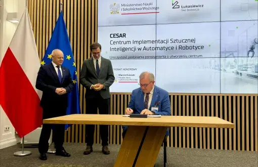 Uroczystość podpisania porozumienia o utworzeniu Centrum Implementacji Sztucznej Inteligencji w Automatyce i Robotyce (CESAR). Profesor Krzysztof Jóźwik siedzi przy drewnianym stole i podpisuje dokument, obok stoją dwaj mężczyźni w garniturach. W tle flagi Polski i Unii Europejskiej oraz ekran z logotypami Ministerstwa Nauki i Szkolnictwa Wyższego oraz Sieci Badawczej Łukasiewicz, a także napisem „CESAR – Centrum Implementacji Sztucznej Inteligencji w Automatyce i Robotyce – Uroczystość podpisania p
