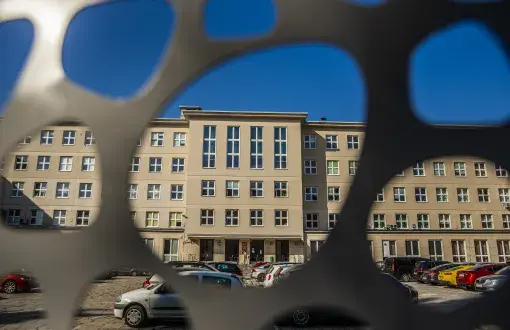 Widok jasnego, kilkupiętrowego budynku z rzędami okien, widziany przez otwory w ciemnym ogrodzeniu. Przed budynkiem stoją zaparkowane samochody, a w tle widać bezchmurne niebieskie niebo.
