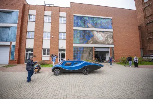 Prezentacja niebiesko-czarnego bolidu solarnego przed ceglanym budynkiem uczelni z mozaiką z luksferów; fotoreporterzy i operatorzy kamer dokumentują pojazd, w tle kilka osób obserwujących wydarzenie.